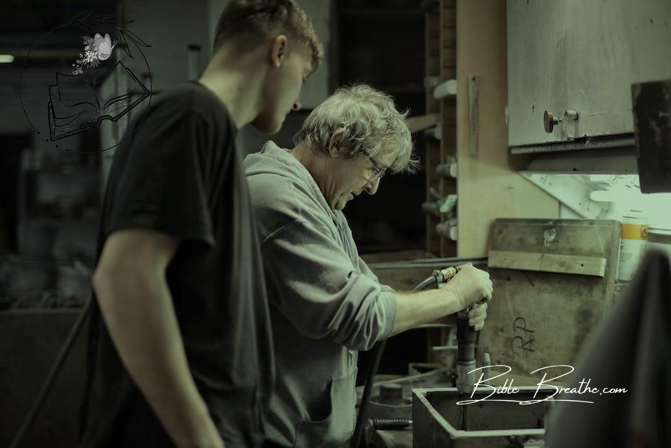 Side view of senior artisan in eyeglasses handling detail in workshop while apprentice looking at process