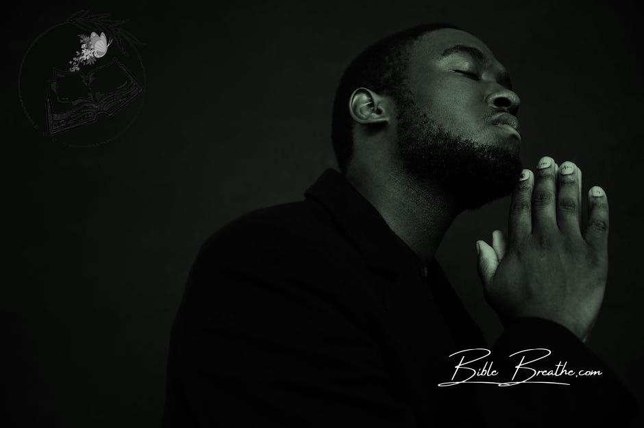 Monochrome handsome unshaven African American male in black suit praying with hands clasped under chin