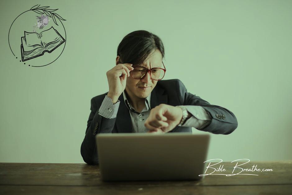 Young frowning man in suit and glasses looking at wristwatch while waiting for appointment sitting at desk with laptop