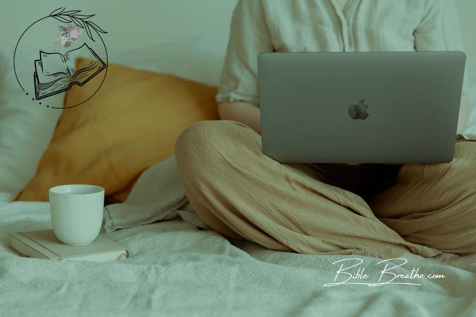 Crop faceless female in casual outfit sitting on bed with legs crossed holding laptop on knees with cup of coffee standing on notebook while working from home
