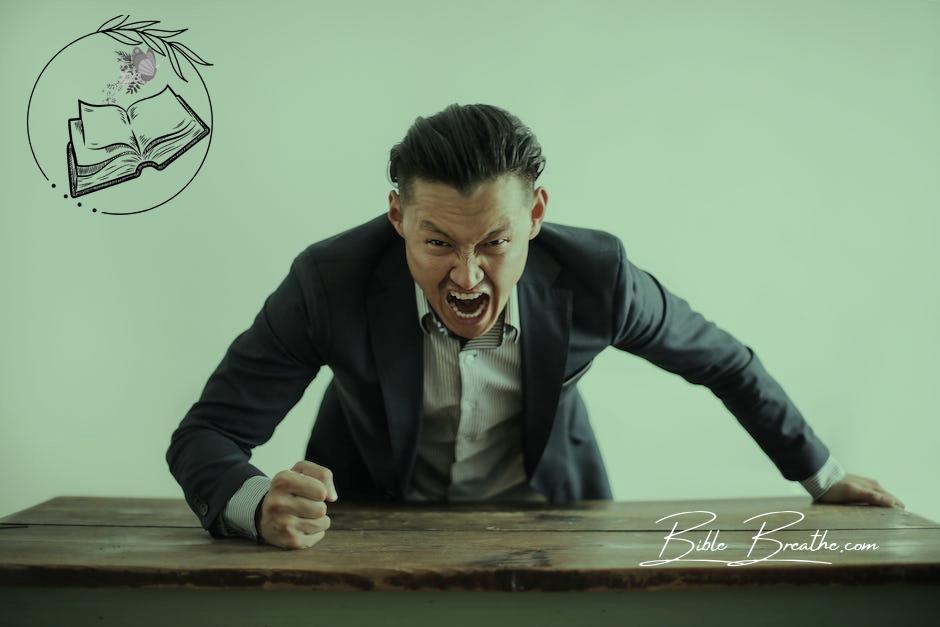 Expressive angry businessman in formal suit looking at camera and screaming with madness while hitting desk with fist