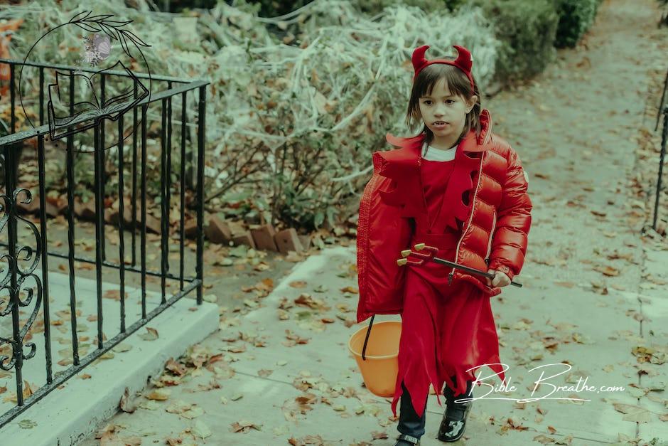 Little girl in red costume of devil for Halloween with horns and pitchfork walking on street in daytime in autumn