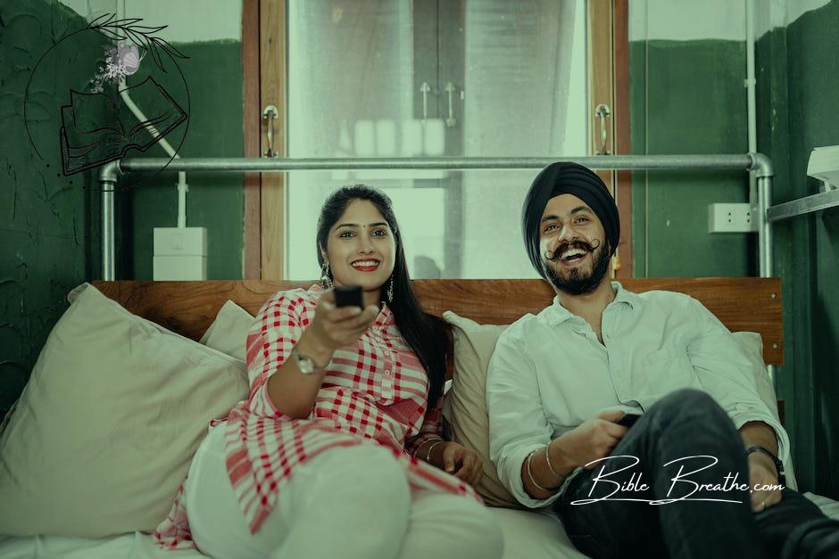 Cheerful wife with bindi on forehead wearing plaid tunic with white trousers using TV remote control for channel switching while lying on bed with laughing Sikh husband in turban with stylish beard and twisted mustache