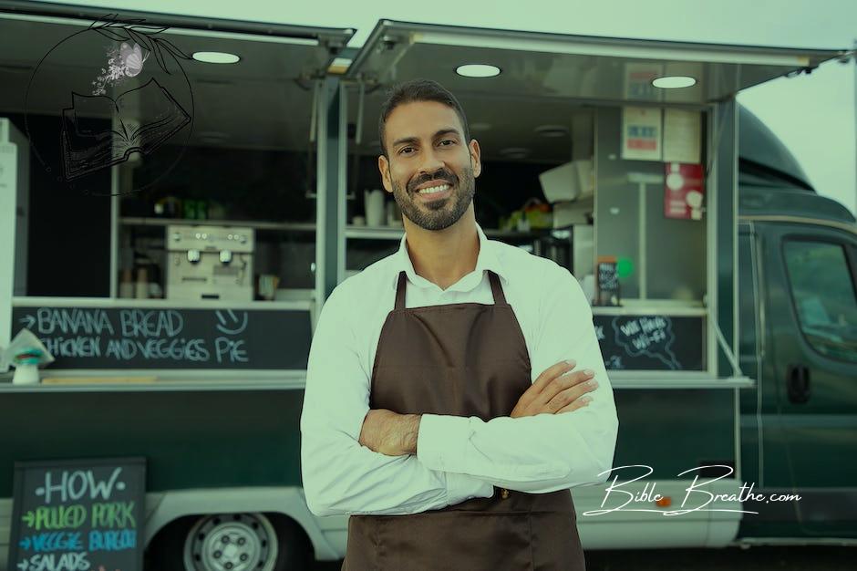 Smiling young ethnic male waiter in apron standing with arms folded near modern food truck and looking at camera contentedly